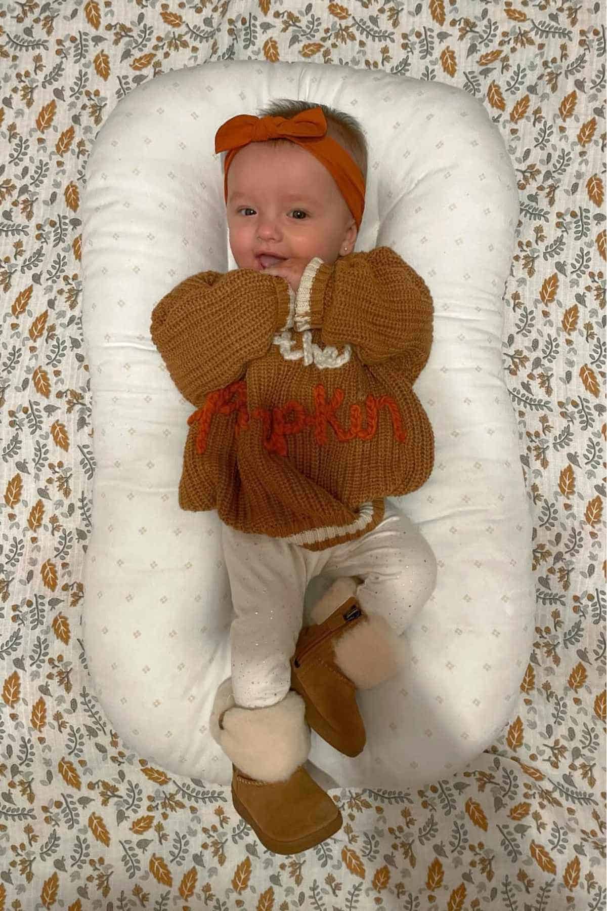 A baby wearing a brown sweater with "Pumpkin" written on it, white leggings, brown boots, and an orange headband, lying on a white pillow with a leaf-patterned blanket underneath.