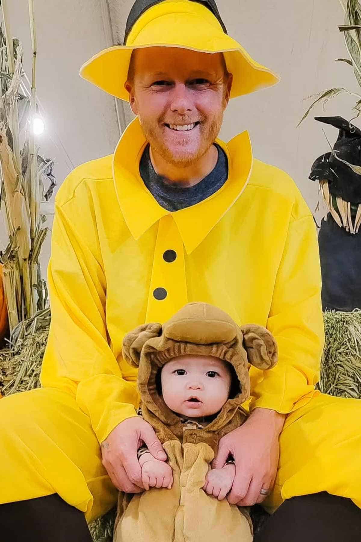 An adult in a yellow raincoat and hat sits behind a baby dressed in a brown monkey costume, both posing indoors on straw bales.