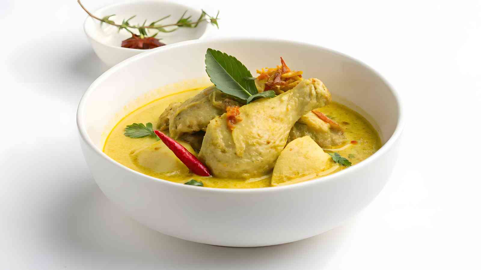A white bowl of yellow curry with chicken drumsticks, potatoes, a whole red chili, and herbs, placed on a white surface with a small bowl of garnish in the background.