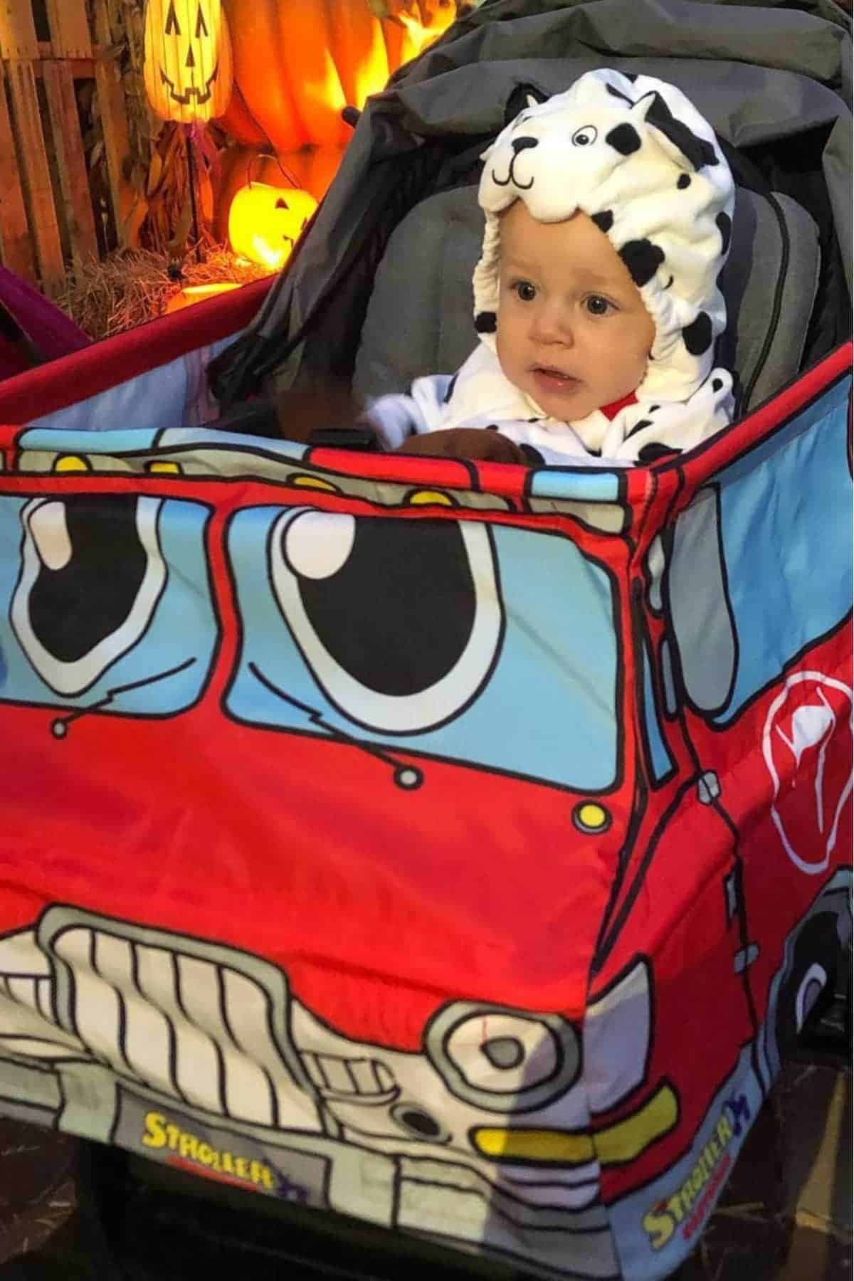 A baby wearing a Dalmatian suit sits in a stroller decorated to look like a red cartoon car, with pumpkins and Halloween decorations in the background.