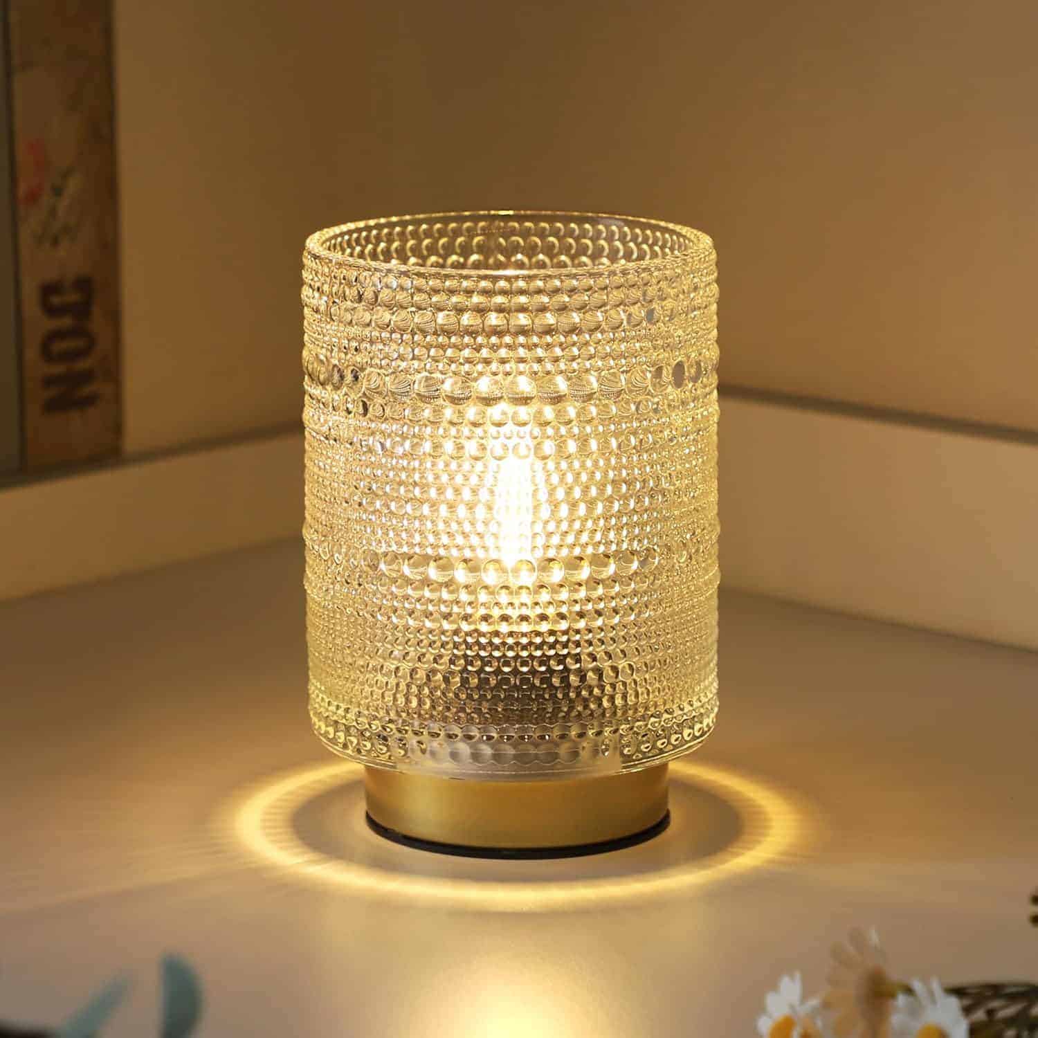 A lit glass table lamp with a textured, beaded surface emits a warm glow, placed on a light-colored surface near a corner.