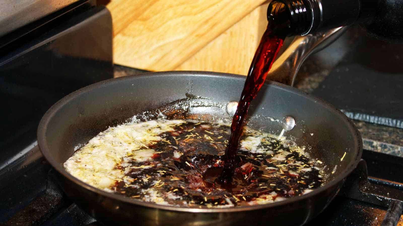 Red wine is being poured into a frying pan with chopped garlic and herbs cooking in oil on a stovetop.