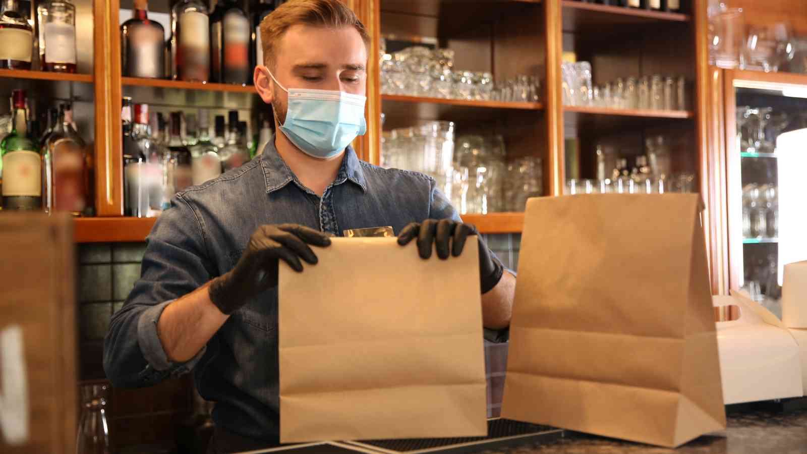 A man wearing a face mask and black gloves packs food into paper bags behind a bar counter lined with glasses and bottles.