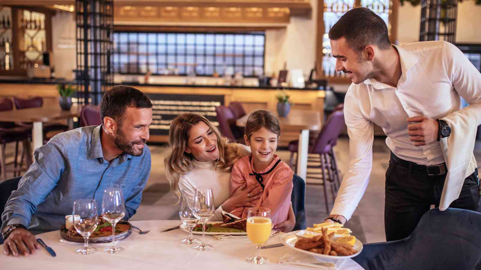 A waiter serves food to a smiling family of three seated at a restaurant table set with drinks and plates.