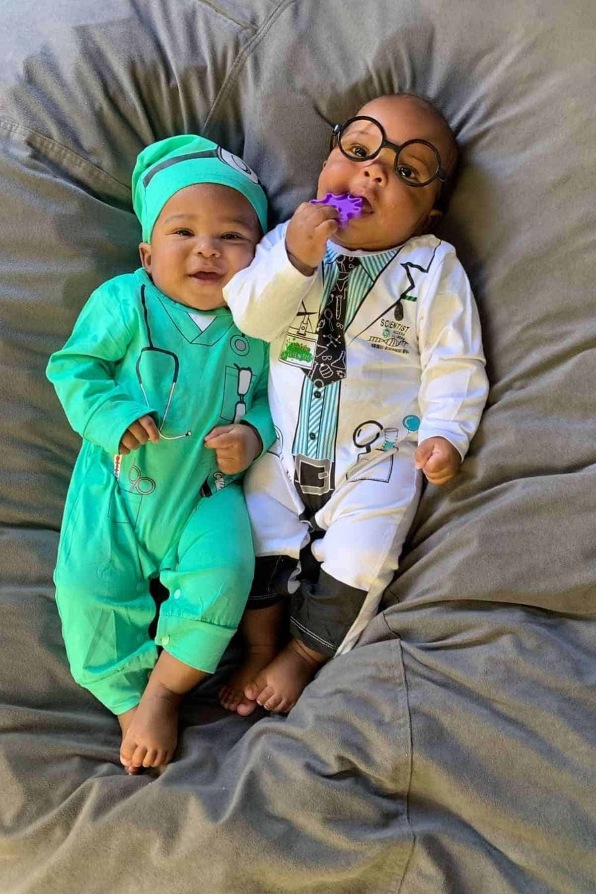 Two babies lie on a gray blanket; one wears a green surgeon outfit and cap, the other wears a white doctor's outfit with glasses and holds a purple toy.