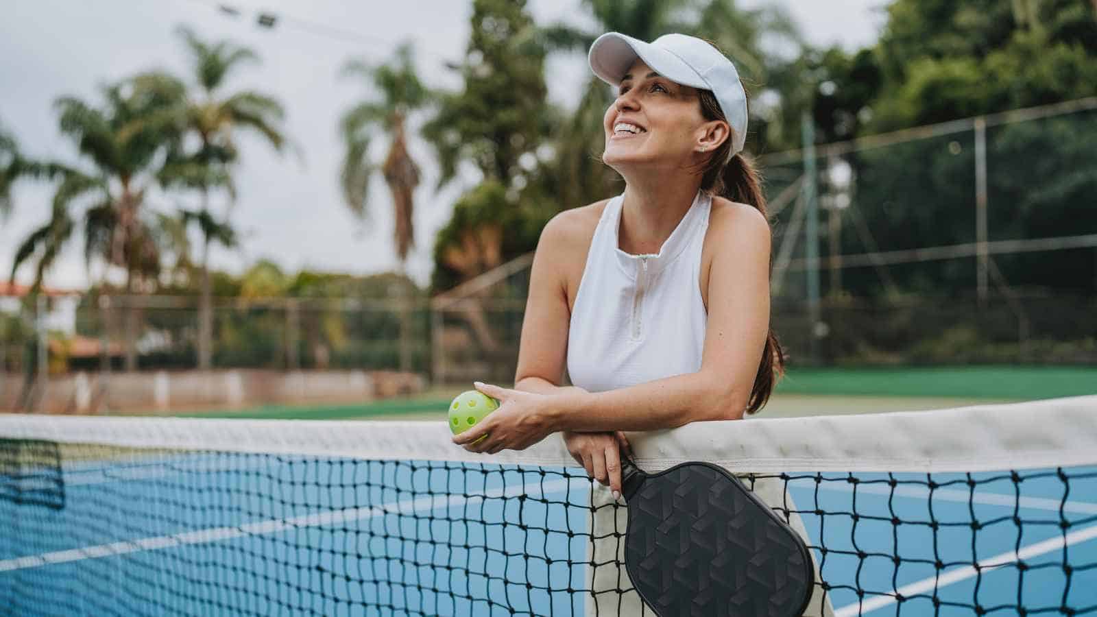 Woman in sports attire holding a pickleball and paddle, leaning on a net on an outdoor court, looking up and smiling.
