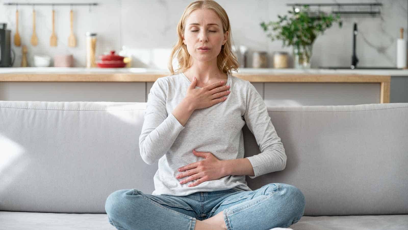 A woman sits cross-legged on a couch with eyes closed, one hand on her chest and one on her abdomen, practicing deep breathing in a kitchen setting.