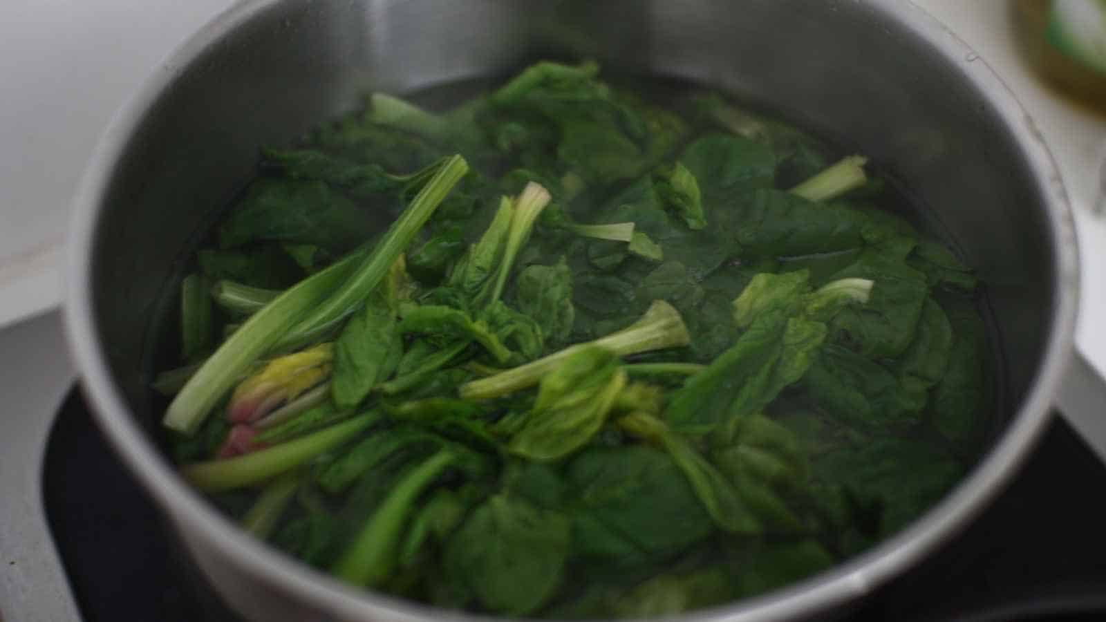 Fresh spinach leaves boiling in a stainless steel pot of hot water on a stove.
