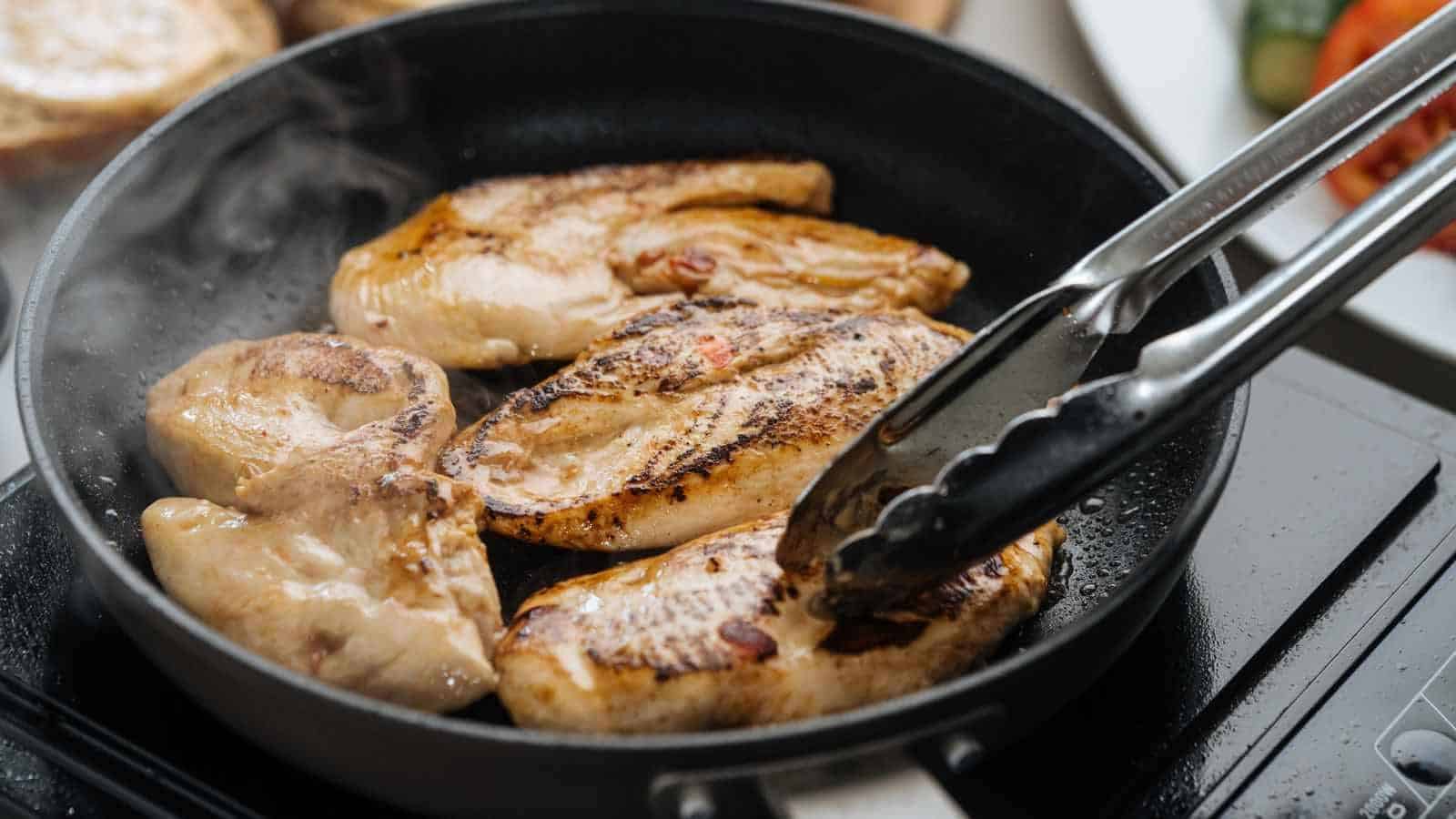 Four pieces of chicken breast cooking in a black frying pan, with metal tongs holding one piece, on a stovetop.