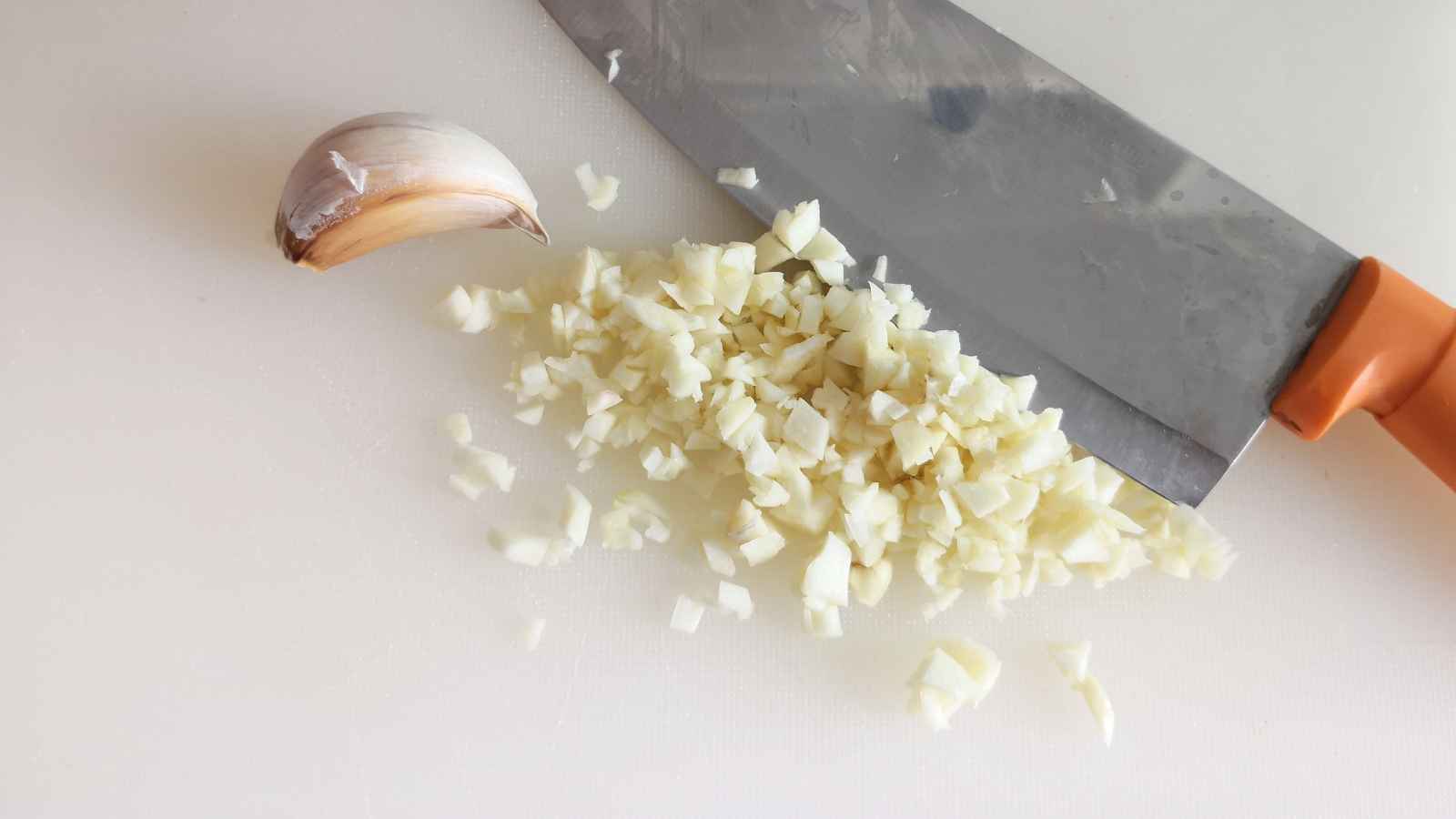 A clove of garlic and a pile of finely chopped garlic are beside a kitchen knife on a white cutting surface.
