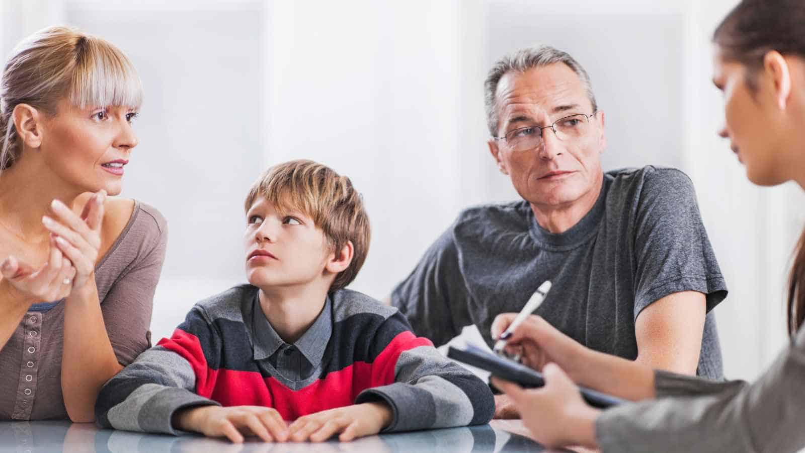 A woman, a man, and a boy sit at a table having a serious conversation with a woman holding a clipboard and pen.