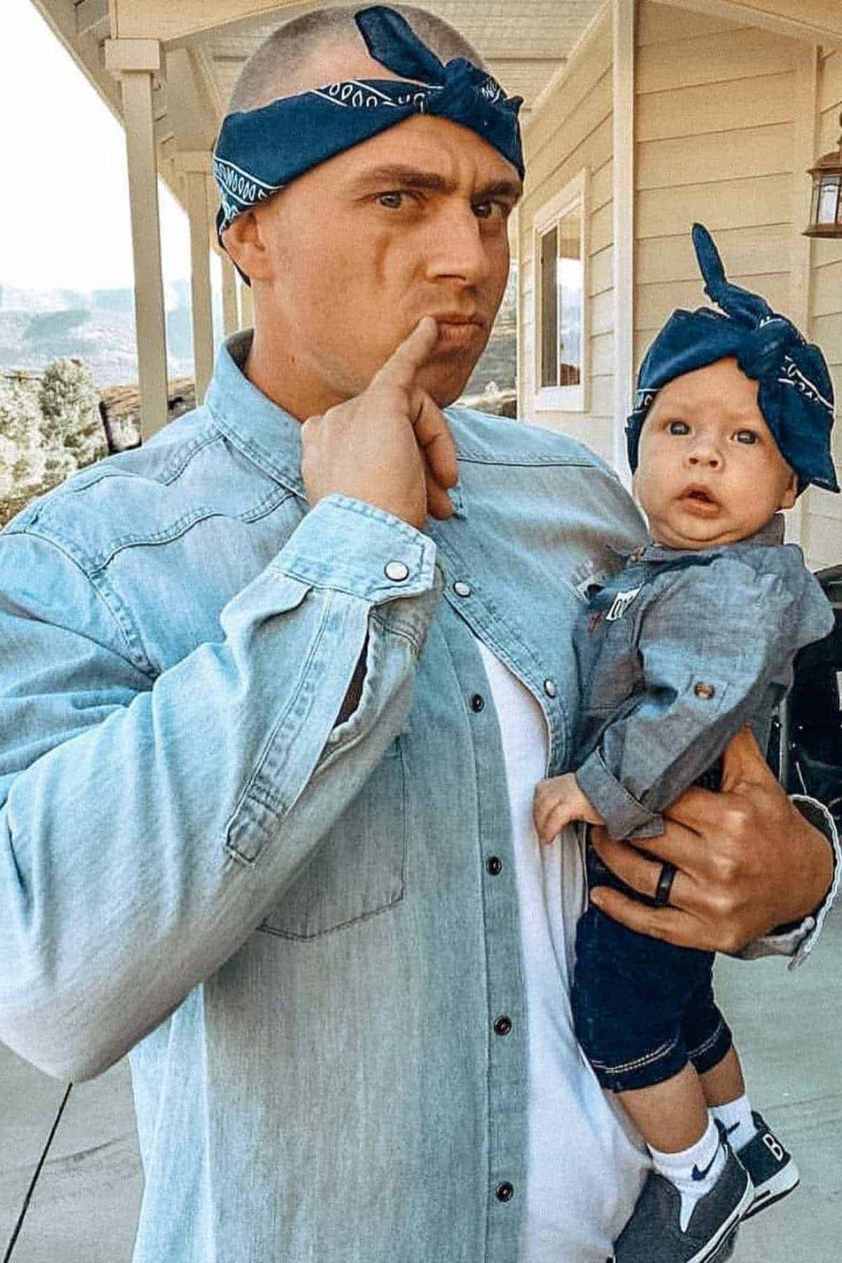 An adult and a baby, both wearing denim shirts and blue bandanas, stand outside a house. The adult is making a thoughtful expression while holding the baby.