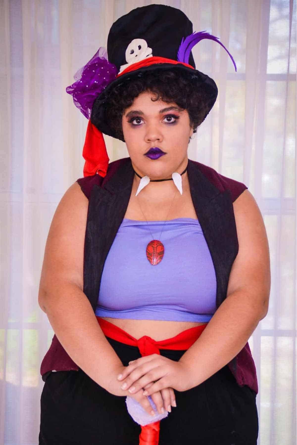 Person in a purple tube top, black vest, and top hat with skull decoration, wearing dark makeup and accessories, standing in front of sheer curtains.