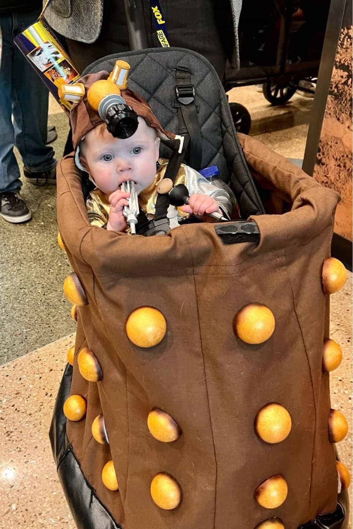 A baby dressed in a Dalek costume sits in a stroller covered with brown fabric and orange bumps, holding a toy.