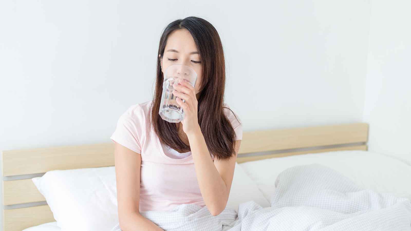 Woman sitting on a bed drinking a glass of water, with white bedding and a light-colored background.