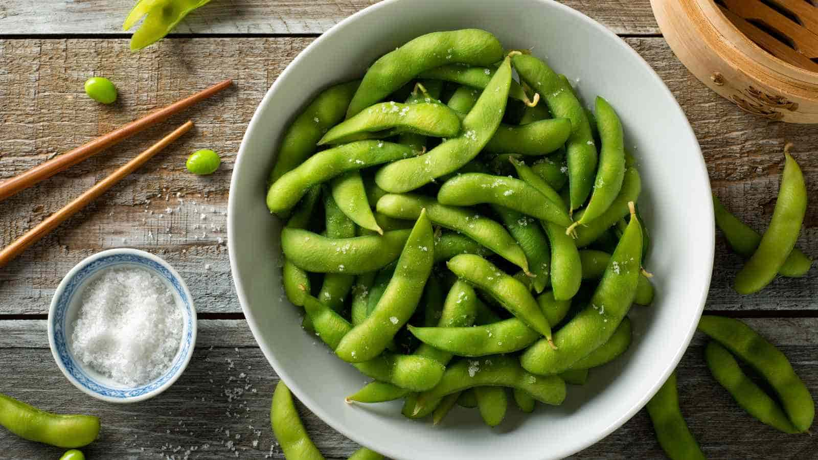 A bowl of salted edamame beans in pods on a wooden table, with chopsticks and a small dish of salt nearby.