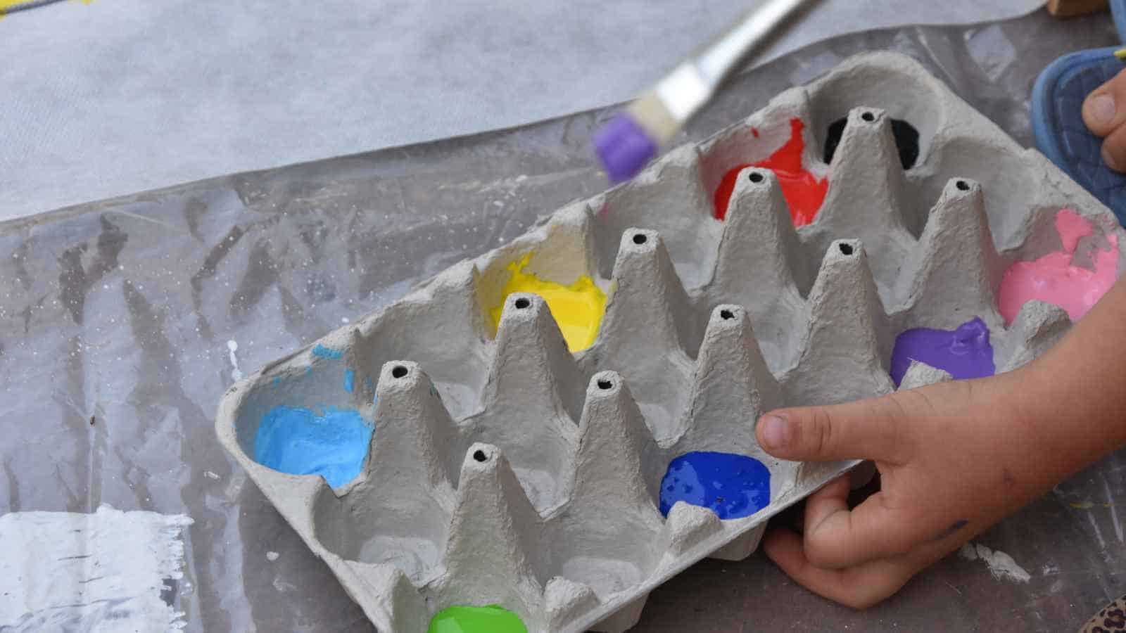 A child's hand holds an egg carton filled with different colors of paint, while a paintbrush with purple paint is visible above the carton.