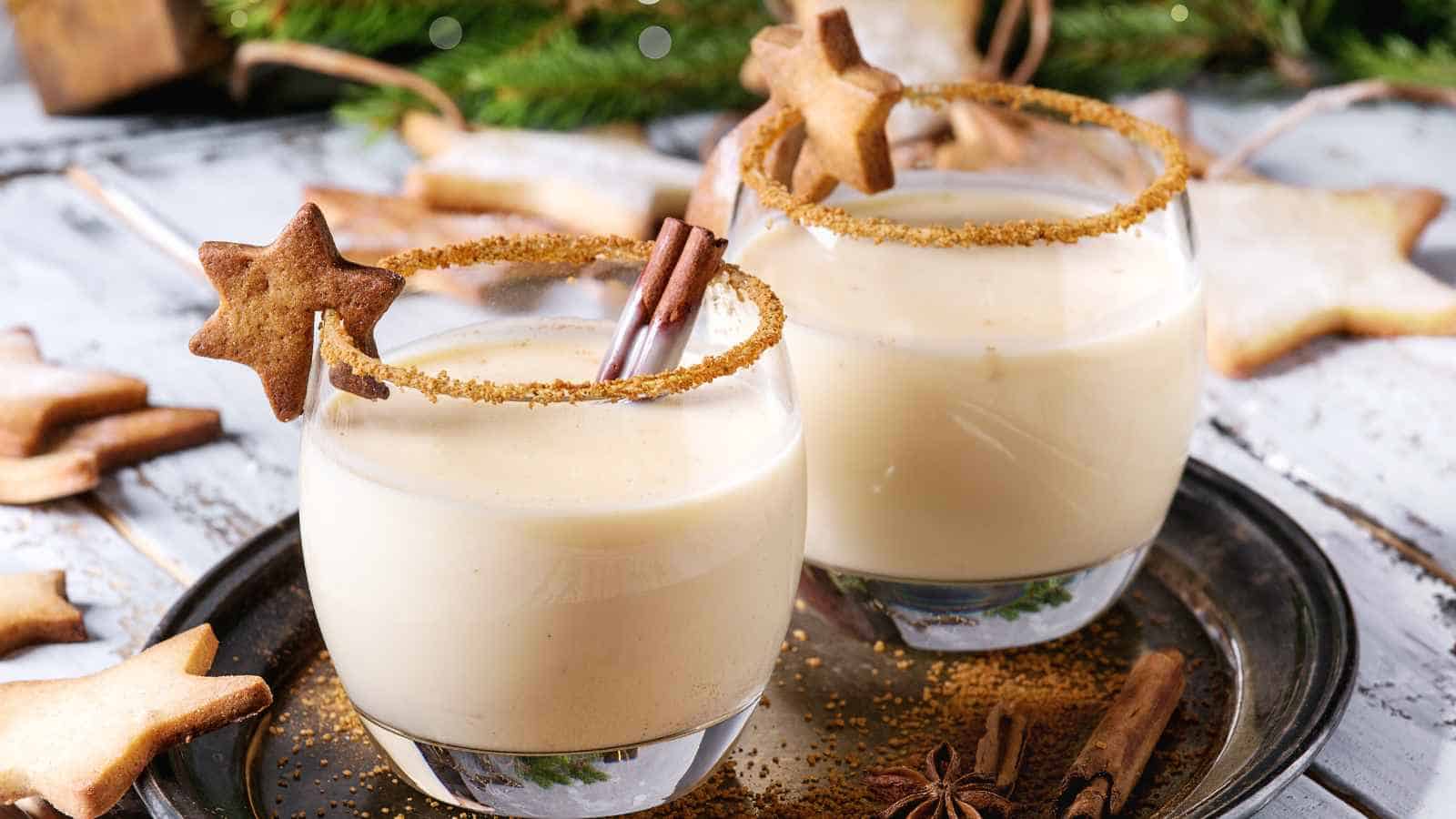 Two glasses of creamy eggnog, each garnished with a cinnamon stick, cookie star, and sugared rim, sit on a tray surrounded by star-shaped cookies.
