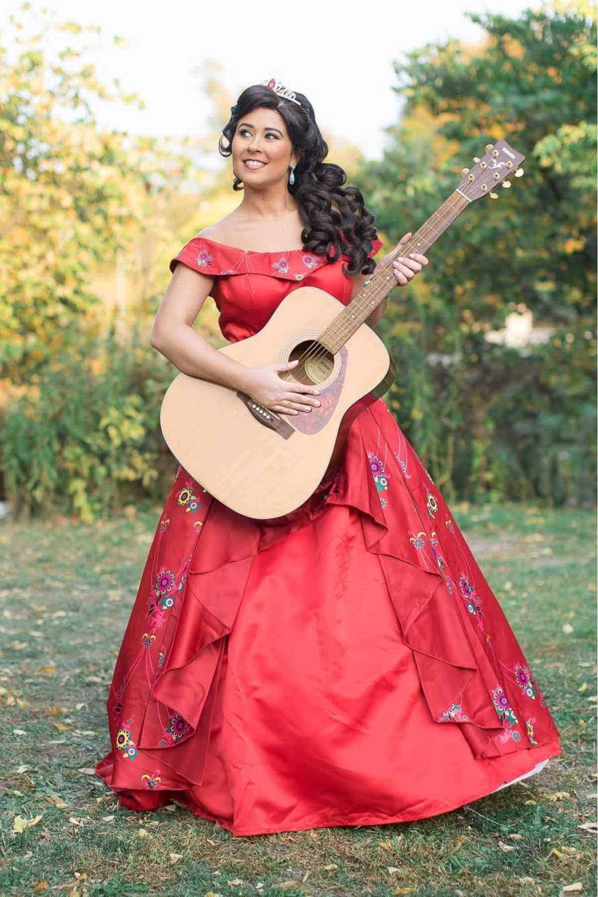 A woman in a red, floral-embroidered gown and tiara stands outdoors, holding and playing an acoustic guitar.
