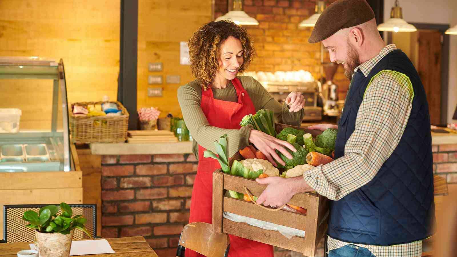 A woman in a red apron receives a wooden crate of fresh vegetables from a man in a hat inside a rustic store or café.