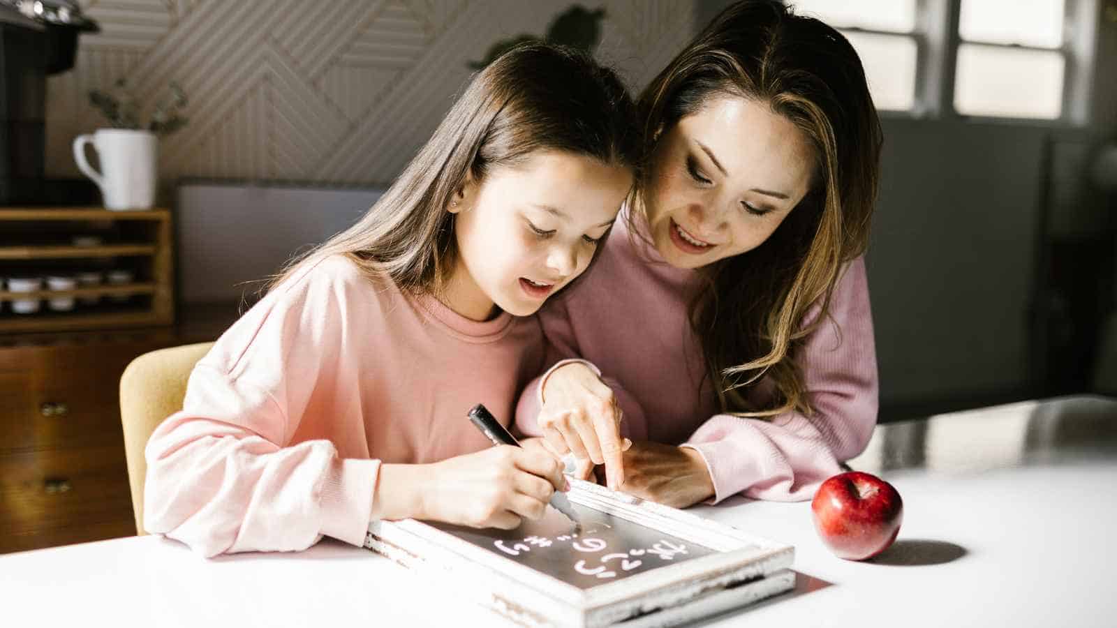 An adult and a child sit at a table, working on math problems written on a whiteboard. An apple is placed on the table beside them.
