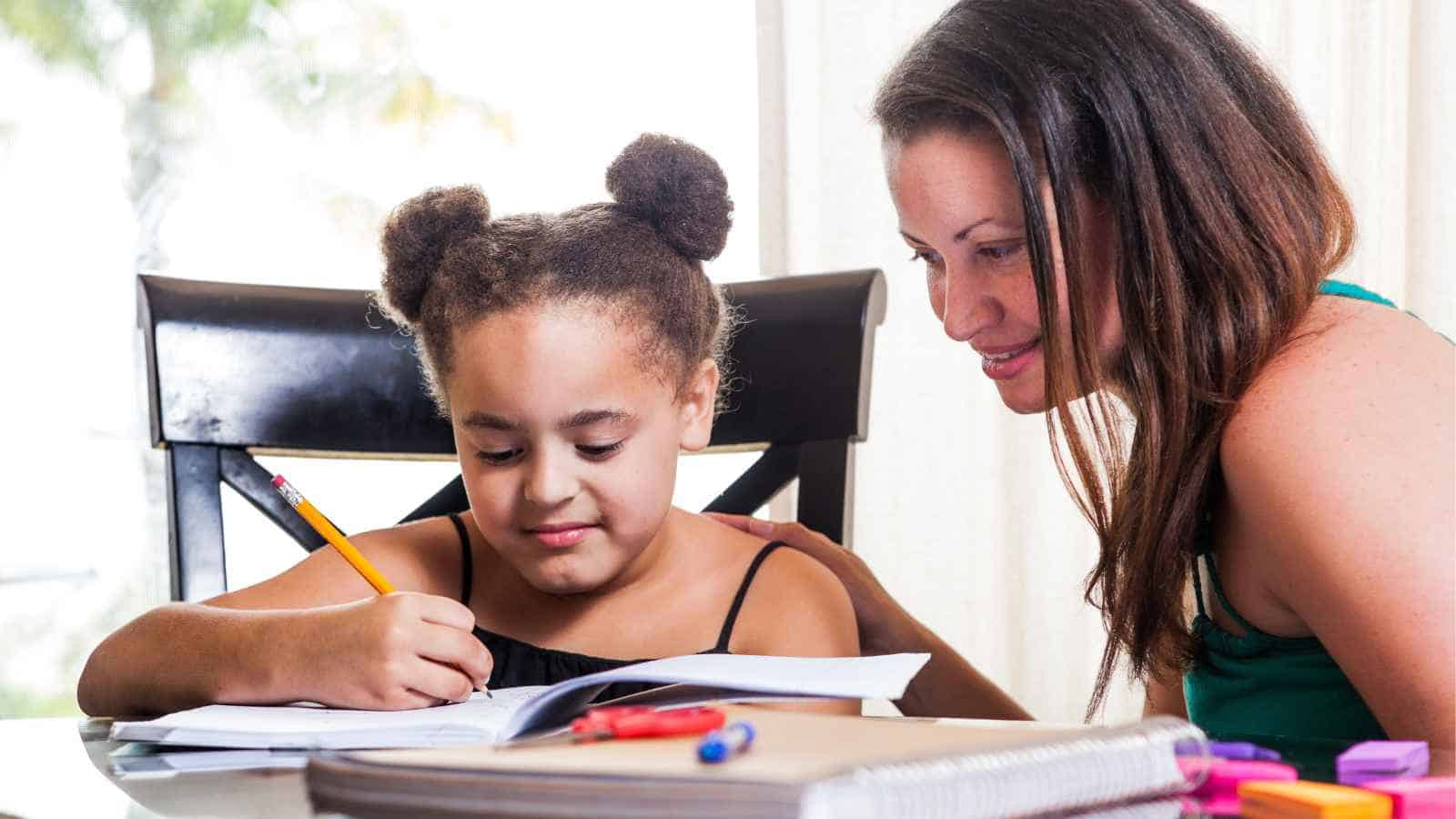 An adult woman helps a young girl with her homework at a table, surrounded by school supplies and notebooks.