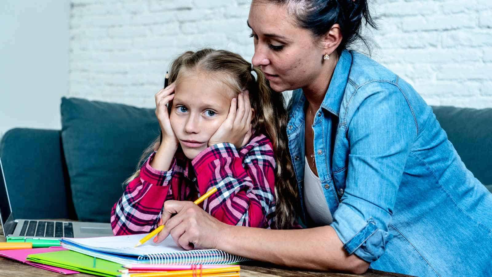 An adult helps a young girl with homework at a table. The girl looks frustrated, resting her head in her hands, while the adult points to her notebook.