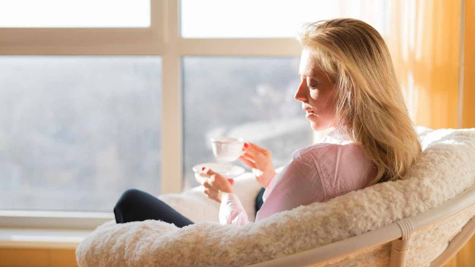 Woman sitting in a white chair by a window, holding a teacup and saucer, with sunlight streaming in.
