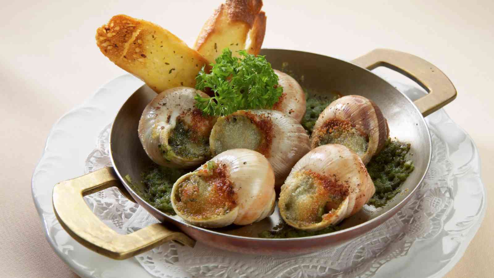 A dish of escargot baked in shells with herb butter, garnished with parsley and served with toasted bread on a metal pan.