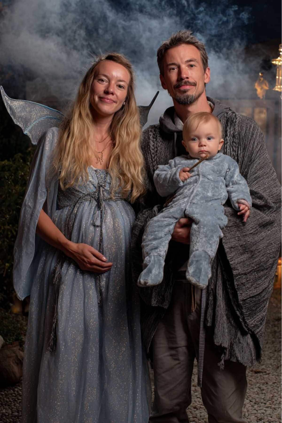A woman in a shimmery dress with fairy wings stands next to a man holding a baby. All are wearing costumes, and there is mist in the background.