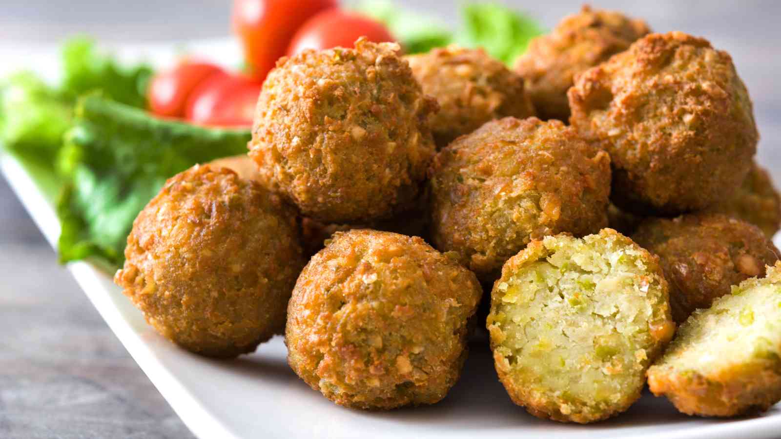 A plate of golden-brown falafel balls with one falafel broken open, showing a green interior, served with lettuce and cherry tomatoes in the background.