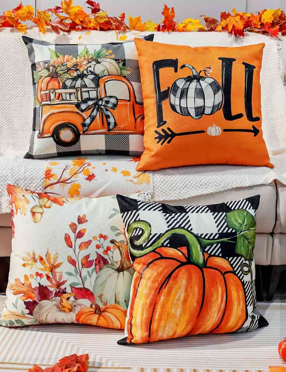 Four decorative fall-themed pillows featuring pumpkins, an orange truck, autumn leaves, and the word "FALL" are arranged on a couch with a garland of autumn leaves above.