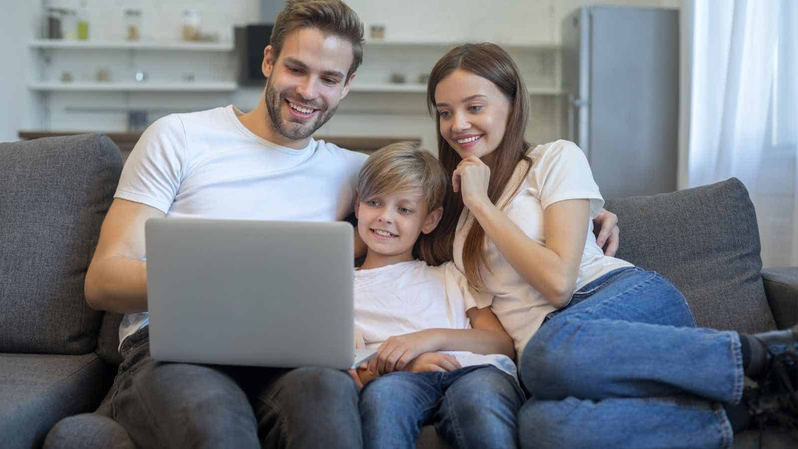 Three people, two adults and a child, sit on a couch together looking at a laptop screen and smiling in a modern living room.
