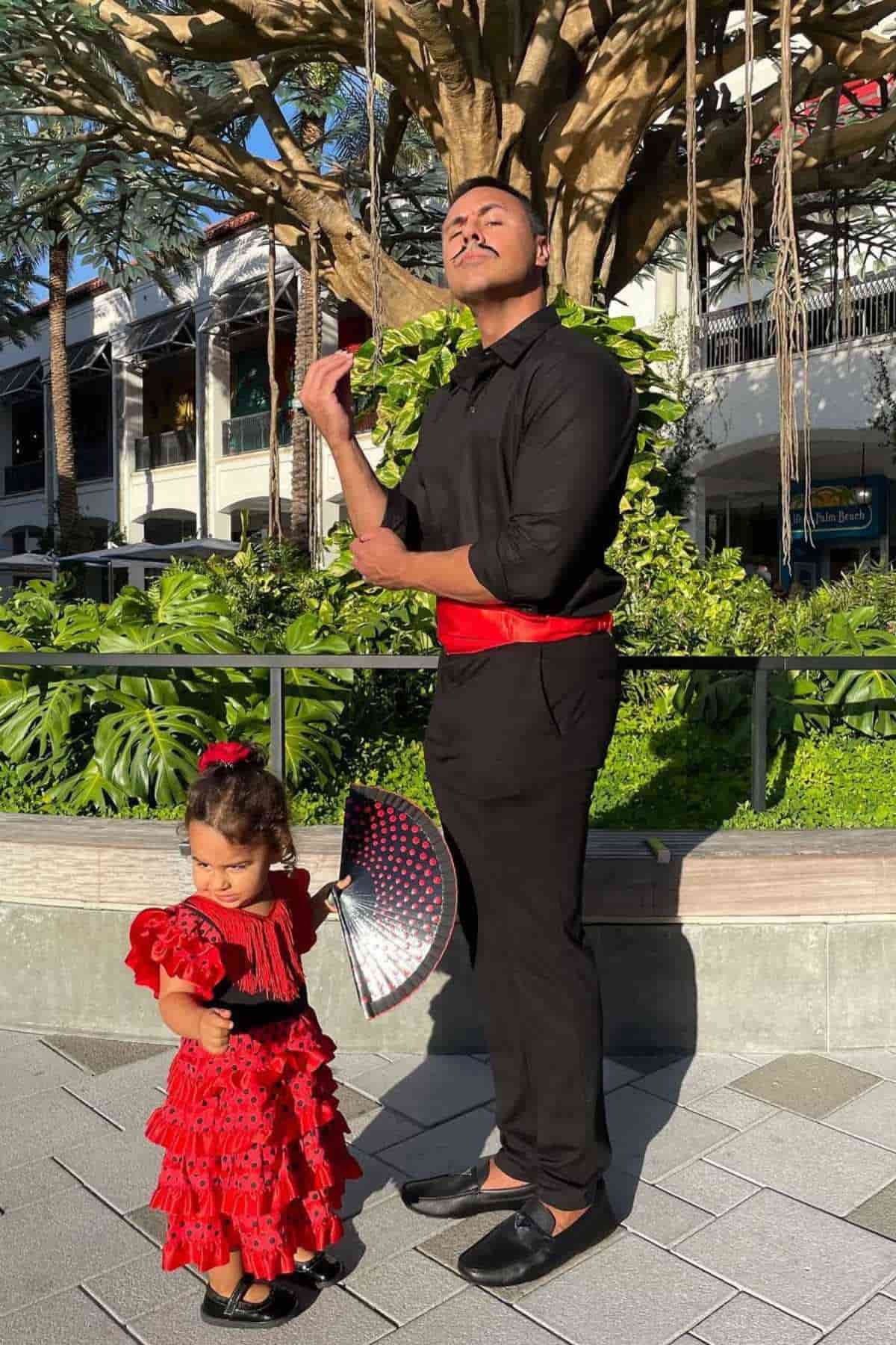 A man and a young girl dressed in matching flamenco-inspired red and black outfits pose outdoors near greenery and a tree.