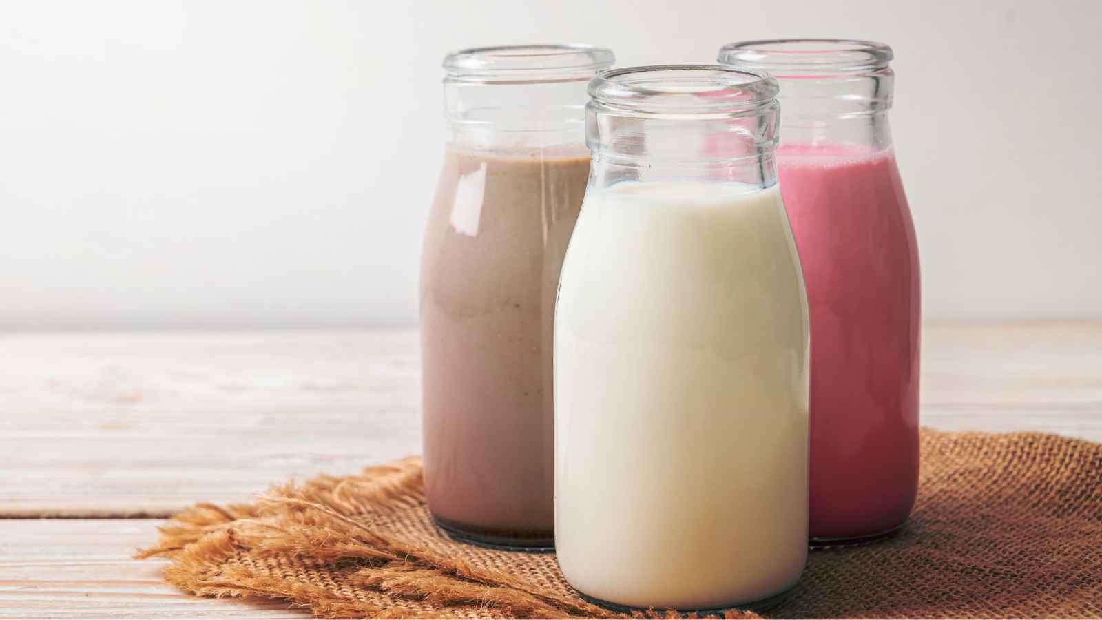 Three glass bottles filled with different types of milk—plain, chocolate, and strawberry—are placed on a burlap mat on a light wooden surface.