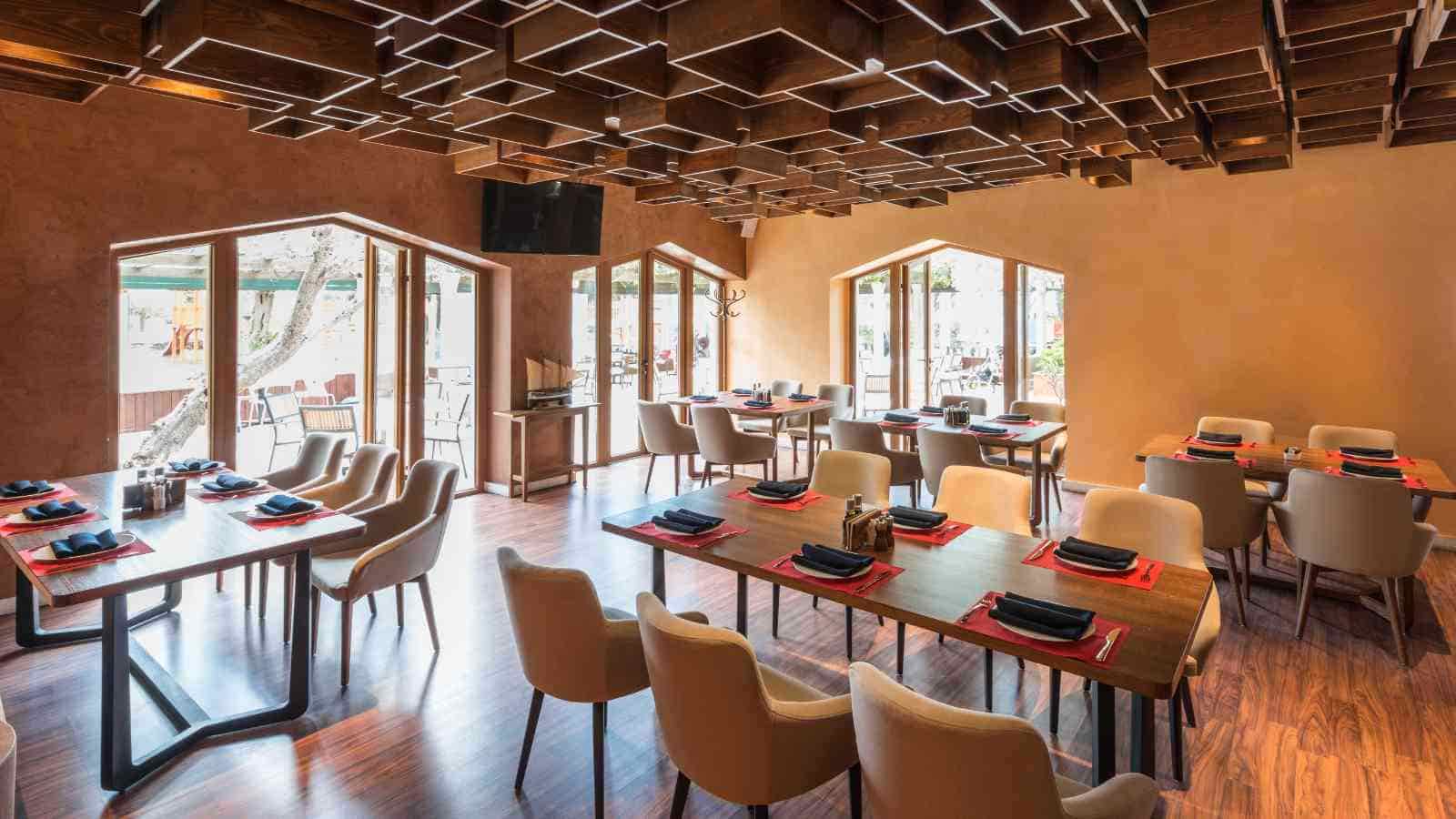 Modern restaurant dining area with wooden tables, beige chairs, place settings, large windows, and a geometric wooden ceiling design.