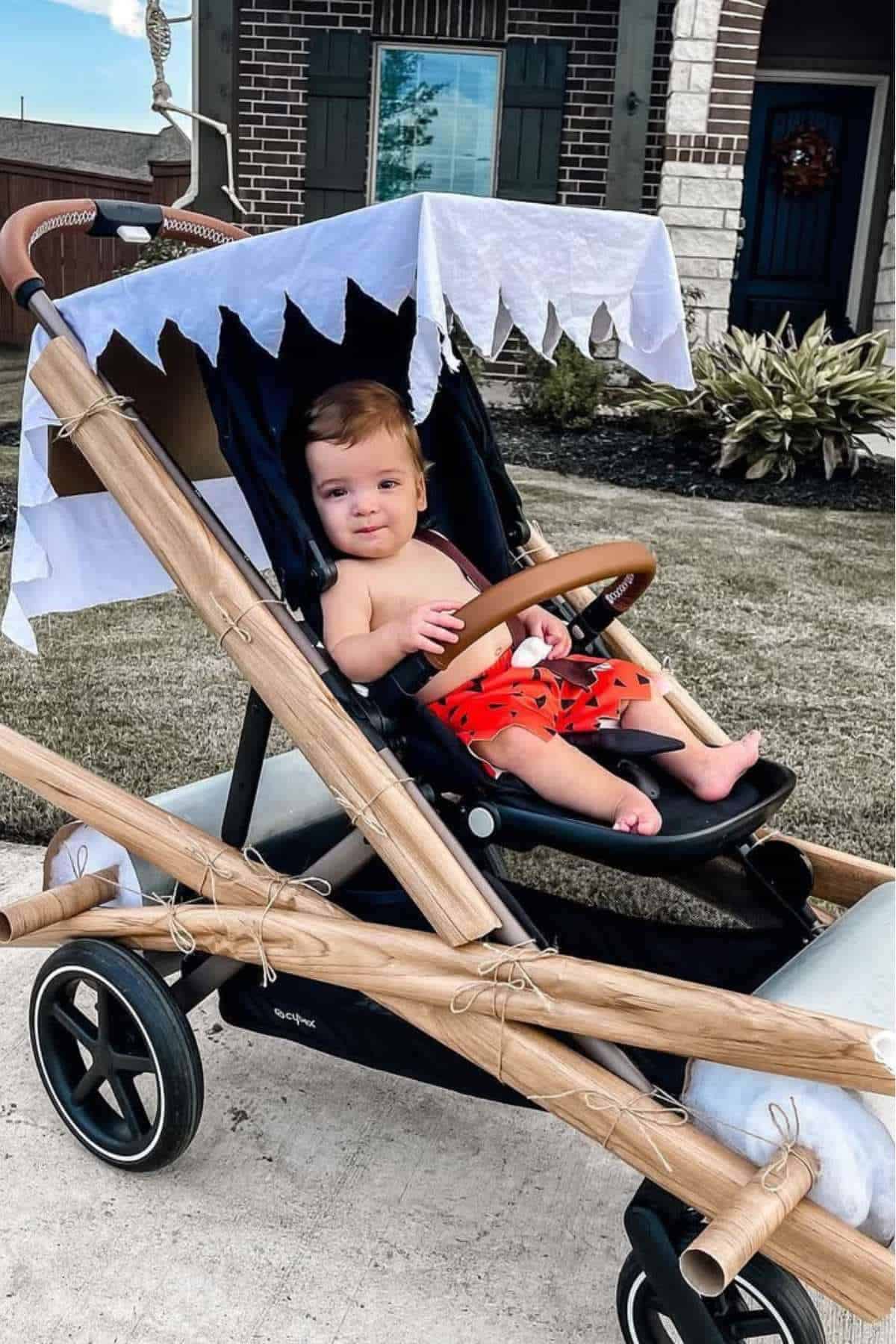 A baby in a stroller decorated to look like the Flintstones' car, with faux wooden poles and a white cloth canopy, sits outside a house.