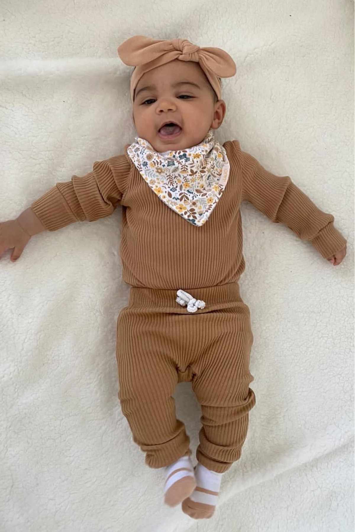 A baby lies on a light blanket wearing a ribbed brown outfit, matching headband, floral bib, and white socks.