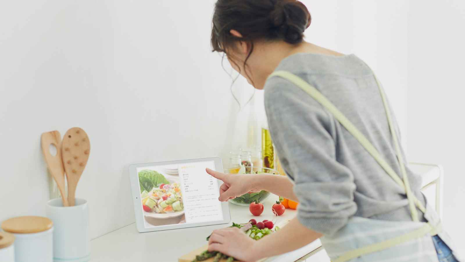 A woman in an apron prepares food in a kitchen while looking at a recipe displayed on a tablet or smart display.