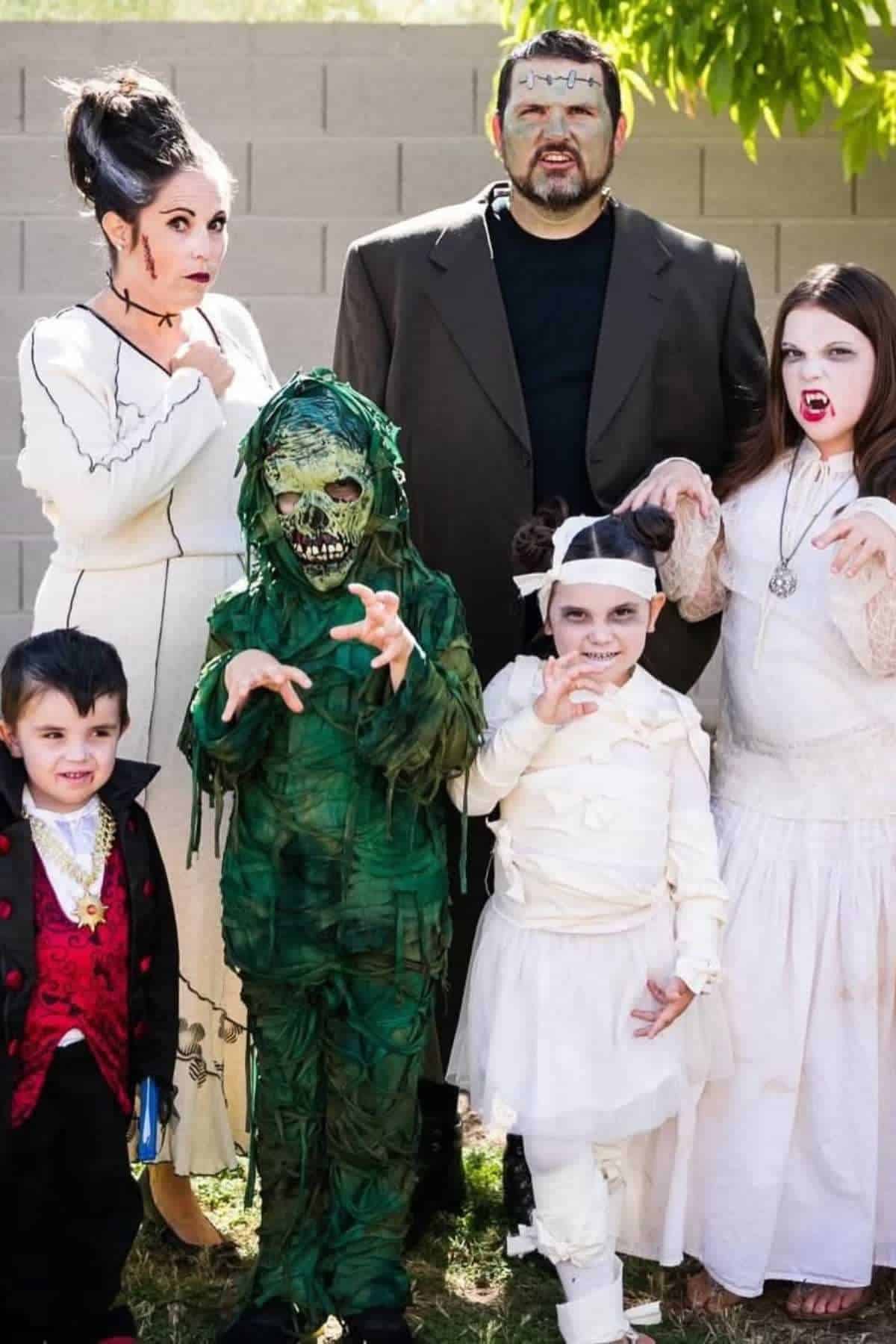 A group of four children and two adults dressed in various classic monster costumes pose outdoors for a photo.
