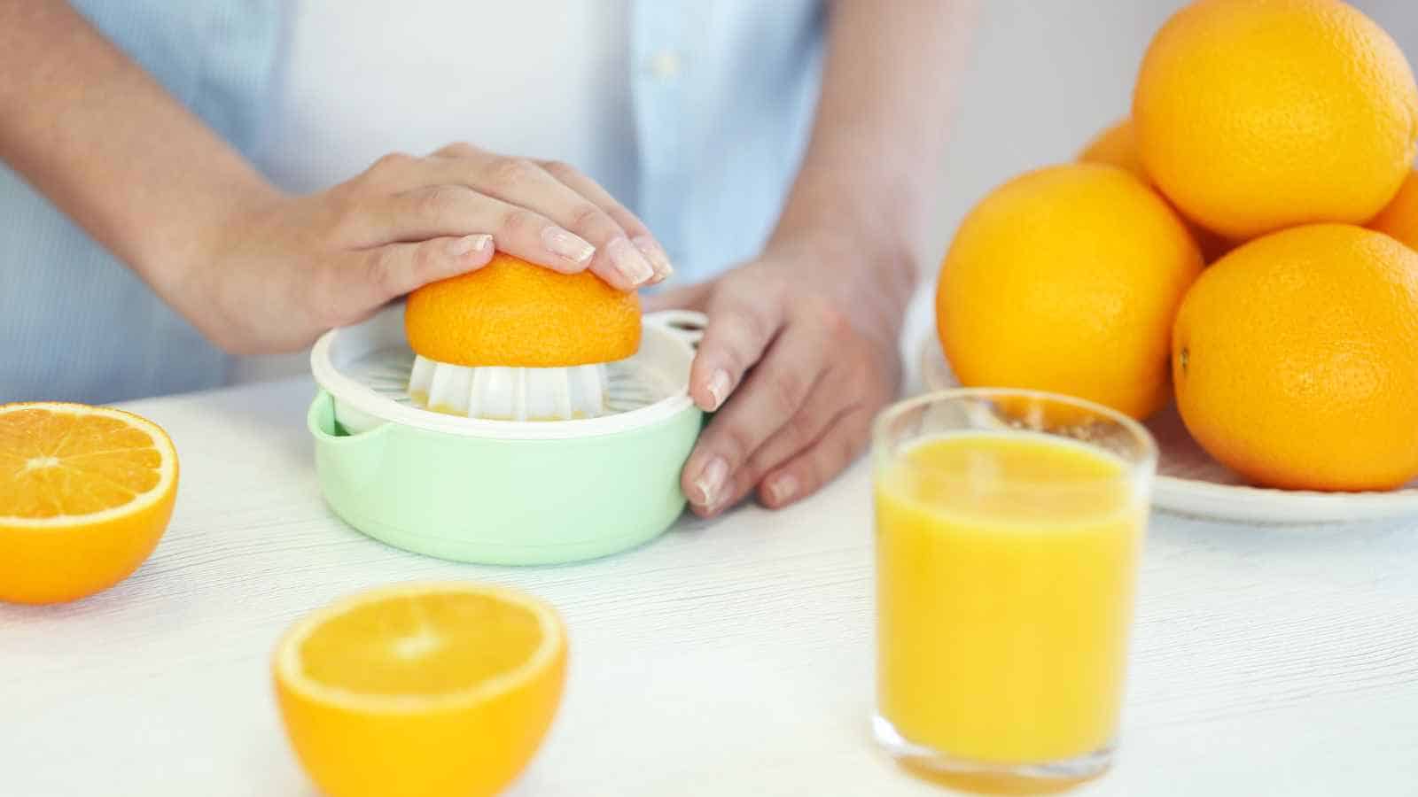 A person presses half an orange on a manual juicer, with a glass of orange juice and whole oranges on a table.