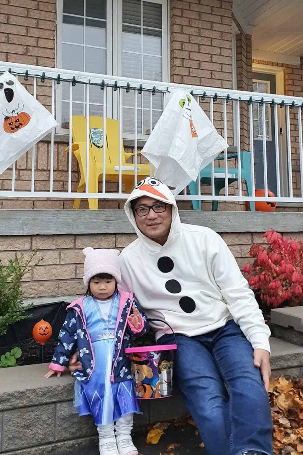 An adult and a child in Frozen-themed costumes sit on a porch decorated with ghost and pumpkin decorations. The child holds a candy bucket.