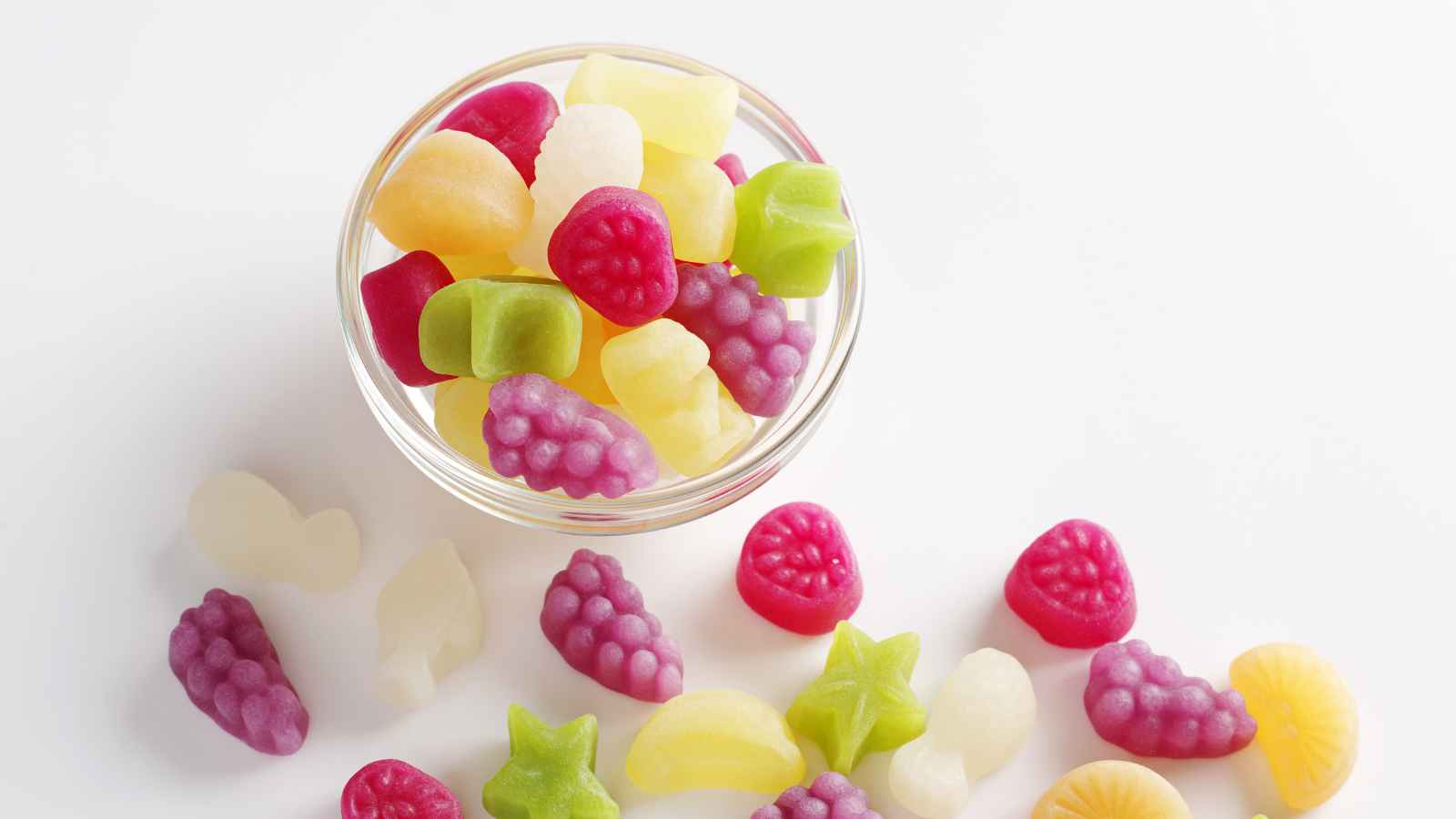 A glass bowl filled with colorful fruit-shaped gummy candies, with additional gummies scattered on a white surface around the bowl.
