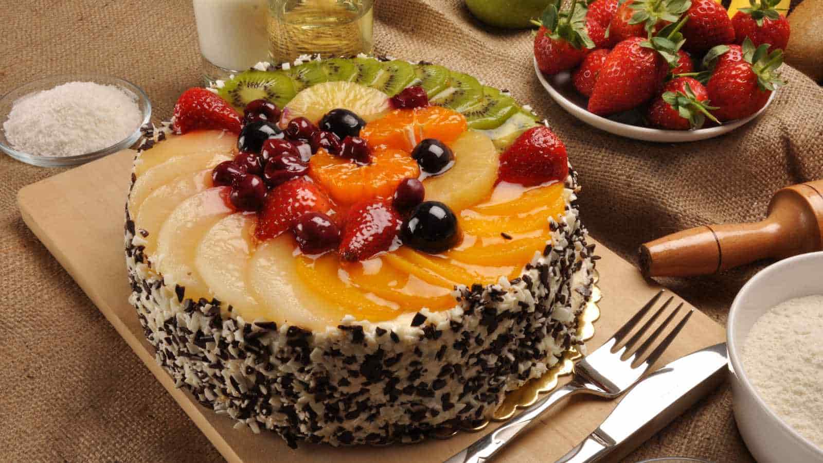 A round cake topped with assorted fresh fruits, surrounded by chocolate shavings, sits on a wooden board with utensils and bowls of ingredients nearby.