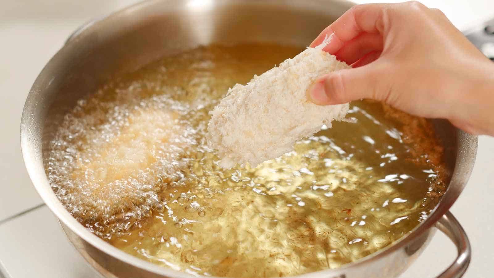 A hand lowers a breadcrumb-coated food item into a pot of hot oil for deep frying.