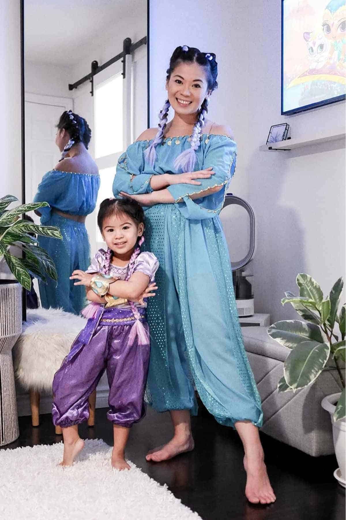 An adult and a child, both wearing blue and purple genie costumes, stand side by side in a home setting with plants and a mirror in the background.