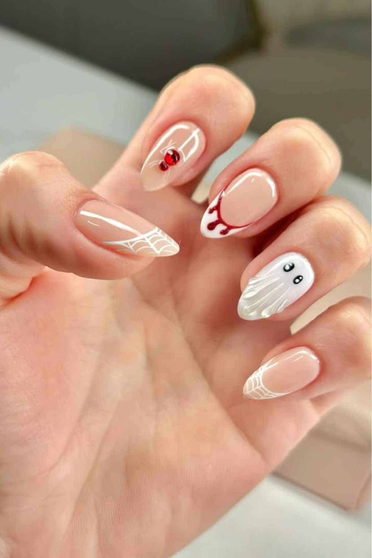 A hand with nude-colored nails features Halloween-themed nail art, including a spider, web designs, blood drips, and a ghost with eyes.