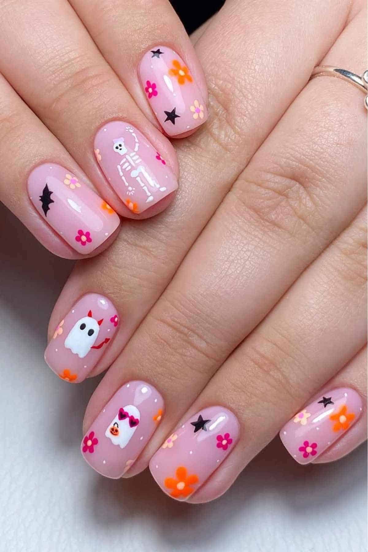 A hand with short nails painted light pink, featuring Halloween-themed designs including a skeleton, ghost, witch, stars, and colorful flowers.