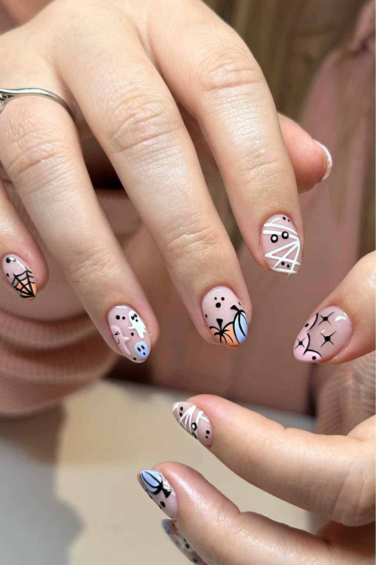 Hands with Halloween-themed nail art featuring designs of ghosts, mummies, spider webs, and bats on a light pink base.