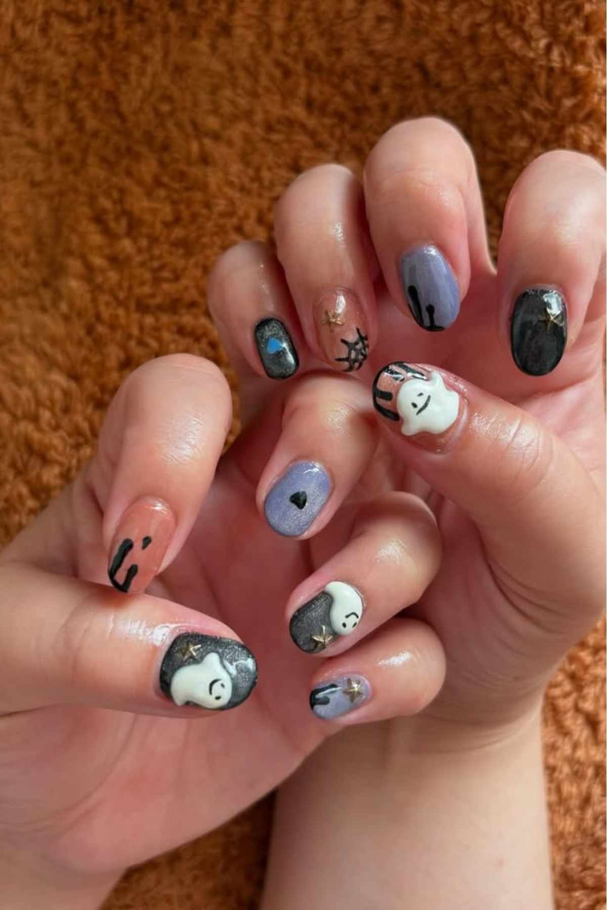 Hands with Halloween-themed nail art featuring ghosts, spider webs, a heart, and black and purple nail polish designs on a brown textured background.